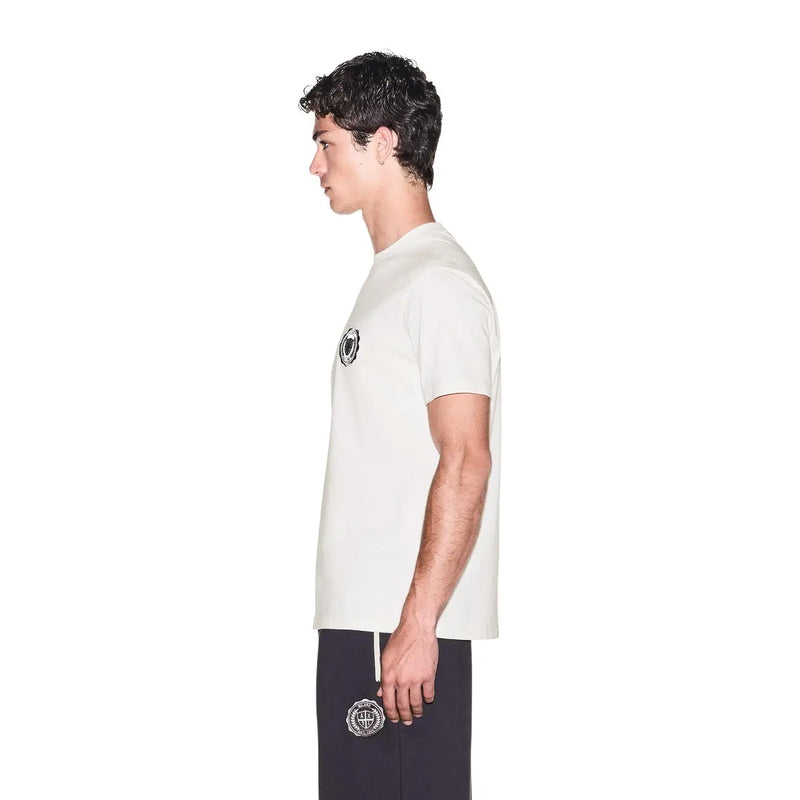 Armani Exchange T-shirt blanc en coton pour homme, manches courtes, logo circulaire noir, porté avec un pantalon bleu marine brodé.