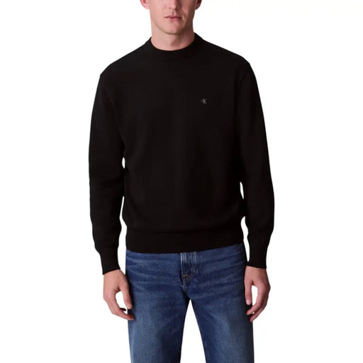 Mannequin homme portant un pull noir à col rond, une chemise grise à manches longues et un pantalon noir.