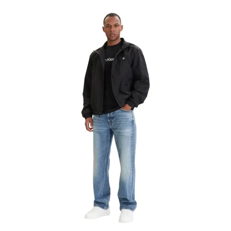 Noir Calvin Klein veste ornée de logos discrets sur la poitrine et la manche, portée sur un ensemble noir assorti. t-shirt avec un jean bleu 100 % coton