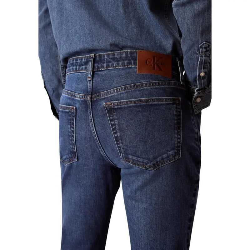 Arrière d'un jean Calvin Klein pour homme en denim de coton bleu avec un écusson de la marque en cuir marron sur la ceinture
