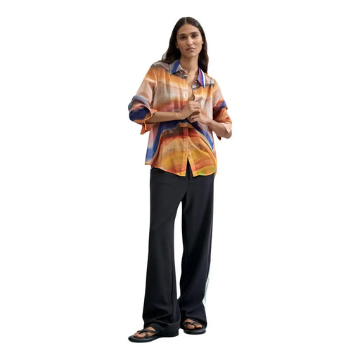 Une femme porte un chemisier Desigual orange en polyester à boutons, orné d'un imprimé abstrait vibrant et de manches fluides.