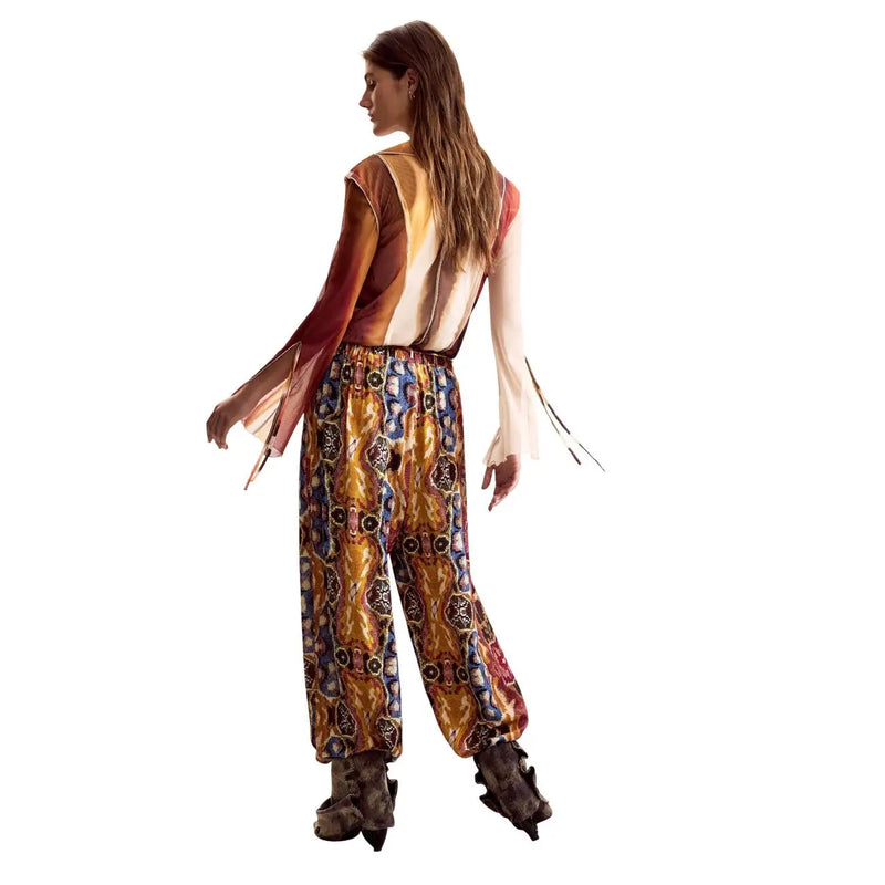Une femme porte un pantalon Desigual moutarde en viscose avec ceinture, associé à une combinaison à motifs colorés.