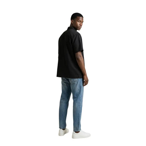 Gianni Lupo Chemise noire à manches courtes pour homme, portée avec un jean bleu et des baskets blanches pour une tenue décontractée.