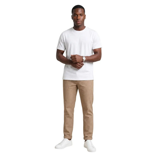 Un homme portant un pantalon en coton beige rayé de la marque Hydra Men, associé à un t-shirt blanc à manches courtes et col rond. t-shirt et des baskets blanches