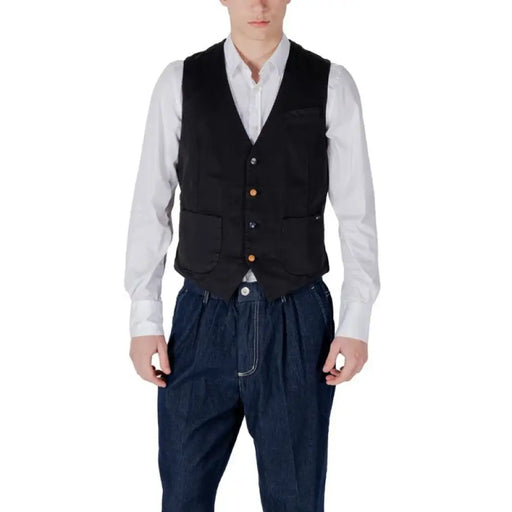Un homme en gilet noir et pantalon bleu présente un gilet bleu marine et une chemise blanche.