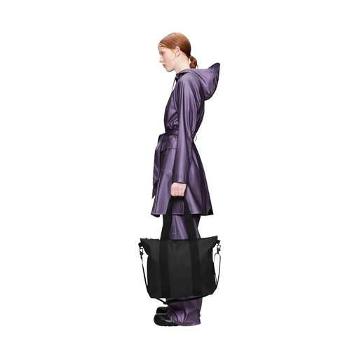 Un élégant mini sac cabas noir Rains pour femme, en polyester brillant avec des anses robustes, porté par un mannequin en imperméable violet.