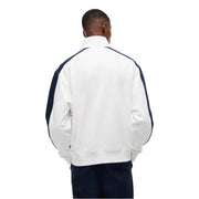 Veste de survêtement Lacoste blanche avec empiècements contrastants bleu marine aux manches et aux épaules, portée avec un jean Tommy Hilfiger et un sweat-shirt en coton bio à rayures blanches.