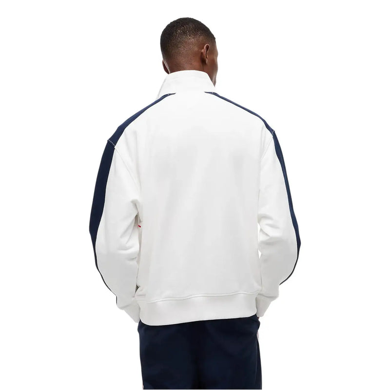 Veste de survêtement Lacoste blanche avec empiècements contrastants bleu marine aux manches et aux épaules, portée avec un jean Tommy Hilfiger et un sweat-shirt en coton bio à rayures blanches.