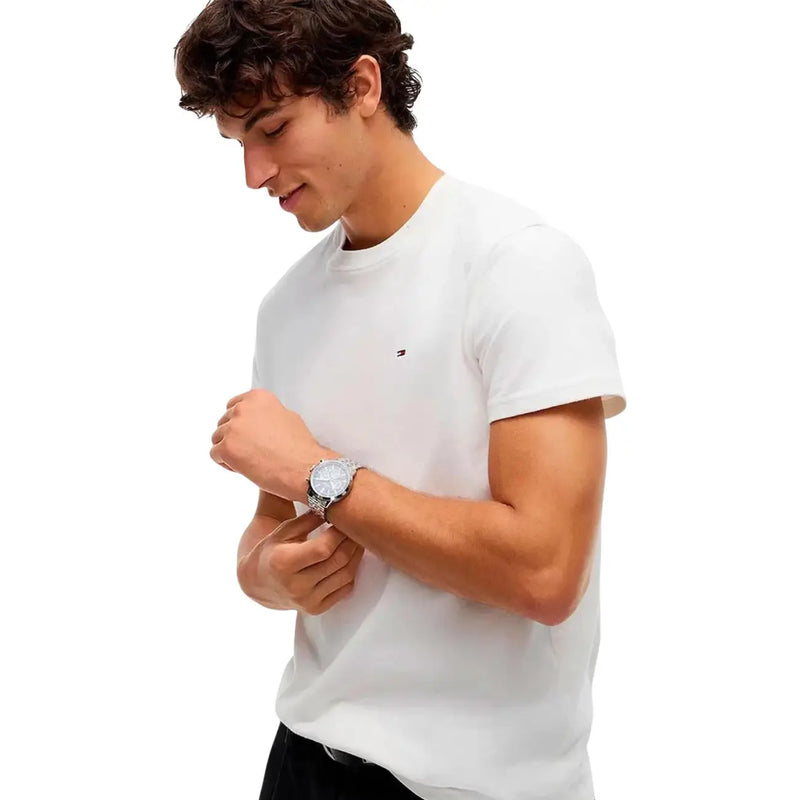 Montre argentée à cadran rond et bracelet métallique portée au poignet avec un t-shirt blanc en coton bio Tommy Hilfiger pour homme