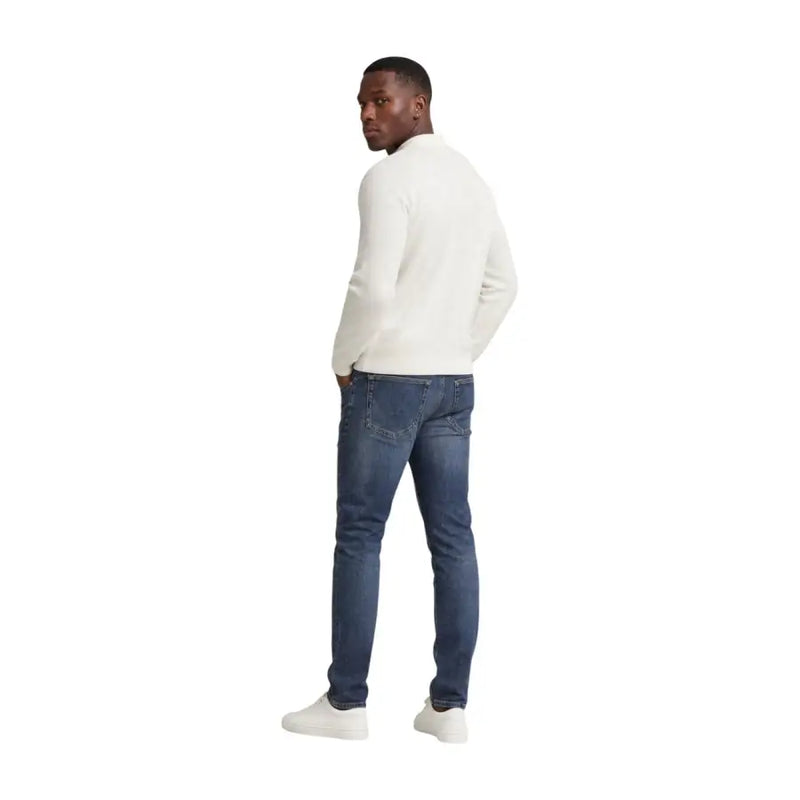 Un homme portant un jean slim bleu en coton Tommy Hilfiger Scanton pour homme, associé à un pull en maille à manches longues couleur crème.