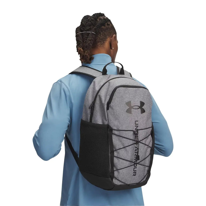 Under armour Sac à dos Hustle Sport 6.0 pour homme, gris, en tissu texturé, avec poche latérale en filet et bretelles croisées.